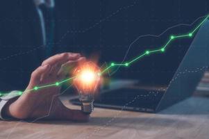 investment strategy and ideas concept, businessman holding light bulb with virtual trading  graph and working on computer laptop on table in office, saving and investment idea, creative new wealth. photo