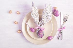 Easter table setting. White plate with a napkin folded in the shape of a rabbit, Easter and chocolate eggs on a pink background. Happy Easter holiday concept. Top view, flat lay photo