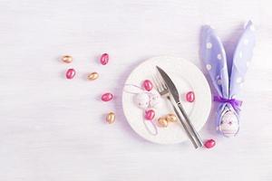 Easter table setting. White plate with a napkin folded in the shape of a rabbit, Easter and chocolate eggs on a pink background. Happy Easter holiday concept. Top view, flat lay photo