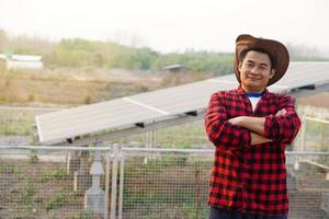 Asian man farmer wears hat, crossed arms on chest, stands beside solar panel at agriculture area, feels confident. Concept, farmer installed photovoltaic for use in agriculture for electricity. photo