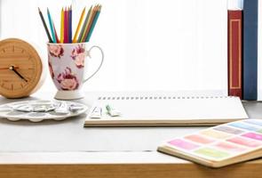 Workspace of watercolor artist with blank sketchbook, palette, color tube, colored pencils, wooden clock and books, side view shot on grey background. photo