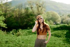 un hermosa mujer en ropa de deporte y un gorra camina en contra un verde natural paisaje y sonrisas en el luz de sol foto