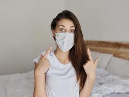 surprised woman in medical mask shows fingers on face and sits on bed photo