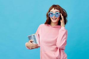 Movie concept. Happy cheerful cute redhead lady in pink hoodie sunglasses with popcorn posing isolated on blue studio background. Copy space Banner. Fashion Cinema. Entertainment offer photo