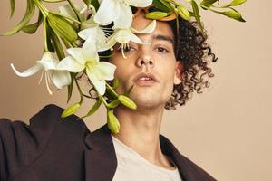 Closeup portrait of handsome tanned curly man in trendy sunglasses hold lilies near face posing isolated on over beige background. Fashion New Collection offer. Retro style concept. Free place for ad photo