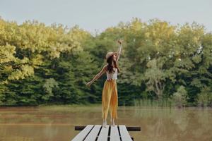 un joven niña en un hippie Mira soportes en un puente por un lago en el otoño y mira a el puesta de sol foto