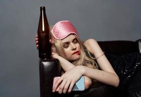 Drunk blonde woman with a bottle of beer sleeps on the sofa indoors on a gray background and a sleep mask on her head photo