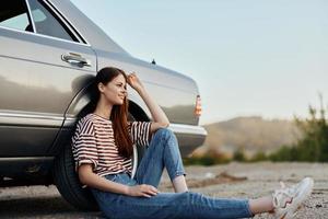 A young woman sits on the ground near her car on the side of the road and looks at the sunset photo