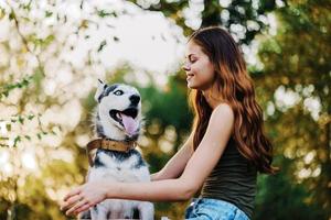 un mujer con un fornido raza perro sonrisas y afectuosamente golpes su amado perro mientras caminando en naturaleza en el parque en otoño en contra el fondo de puesta de sol foto