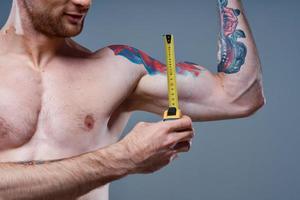 guy measures arm muscles with centimeters on a gray background and a multicolored tattoo bodybuilder fitness photo