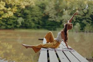 un hippie mujer se sienta en un puente en el banco de un río y disfruta el hermosa paisaje alrededor su, contento en el puesta de sol otoño Dom. ecológico estilo de vida foto