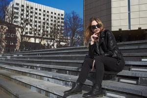 Woman in leather jacket sits on the stairs near the building in the autumn on the street photo