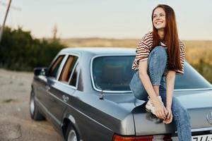 A young woman sits on the trunk of a car and rests after a difficult road and admires nature with a beautiful view. Stopping is also part of the journey photo