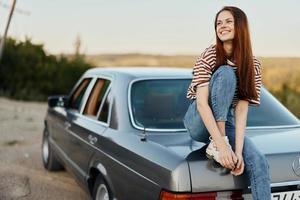 un joven mujer se sienta en el maletero de un coche y descansa después un difícil la carretera y admira naturaleza con un hermosa vista. parada es además parte de el viaje foto