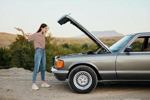 un mujer abrió el capucha de un roto abajo coche y intentos a encontrar el porque de el Descompostura en el la carretera durante un naturaleza viaje solo. foto