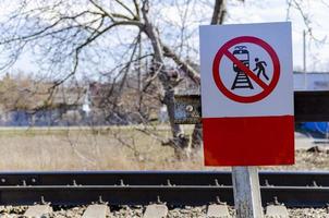 A red sign with a black silhouette of a person stepping on the railway tracks, which prohibits passengers or other people from crossing the railway tracks. Square sign, background of trees, safety on photo