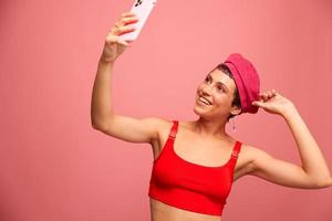 A young woman blogger with colored pink hair and a short haircut takes a picture of herself on the phone and broadcasts a smile in stylish clothes and a hat on a pink background monochrome style photo