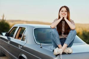 A young woman sits on the hood of her car and laughs with her eyes closed, a journey from a noisy city to nature in summer, Travel lifestyle photo