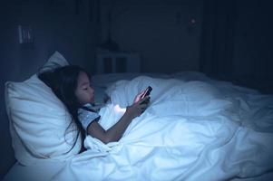 asiático niño jugando juego en teléfono inteligente en el cama a noche, la niña adicto social medios de comunicación foto
