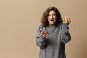 Excited happy curly beautiful lady in gray casual sweater get cool news from message posing isolated on over beige pastel background. Social media, network, distance communication concept. Copy space photo