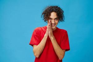 Sly curly tanned Caucasian guy in basic red t-shirt rubs his hands posing isolated on over blue background. Lifestyle and Emotions concept. Good offer with copy space for ad photo