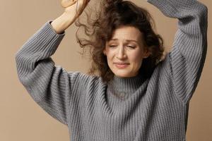 Tortured unhappy curly beautiful female in gray casual sweater with hairbrush can't comb her hair posing isolated on over beige pastel background. Problematic unruly damaged hair concept. Copy space photo