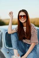 A fashionable woman smiles sweetly in stylish sunglasses, a striped t-shirt and jeans, sits on the trunk of a car and looks at the beautiful nature of autumn. Travel lifestyle photo