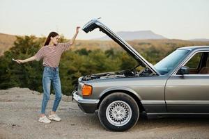 A woman opens the hood of a broken down car and tries to find the cause of the breakdown on the road during a nature trip alone and is sad photo