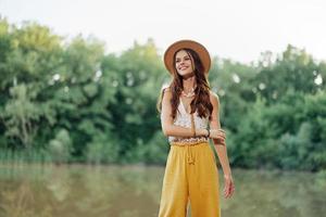 un joven mujer en un hippie Mira viajes en naturaleza por el lago vistiendo un sombrero y amarillo pantalones en el otoño foto