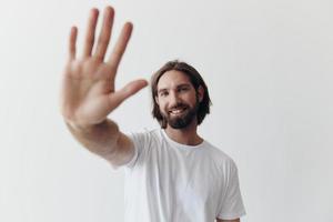 Happy adult man with a beard smiles and pulls the cook into the camera listening to music in headphones in a distressed t-shirt on a white isolated background photo