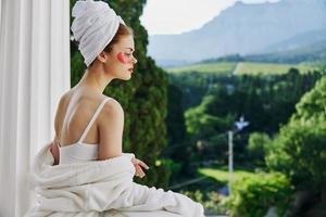 mujer en un bata de baño en un blanco túnica el balcón pasa por alto el montañas mirando en el ver foto