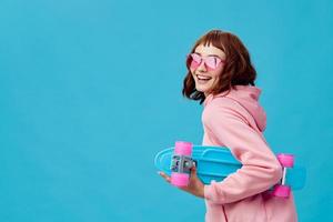 Funny joyful pretty redhead lady in pink hoodie sunglasses with penny board smiling posing isolated on blue studio background. Copy space Banner Offer. Fashion Cinema. Holiday activity photo