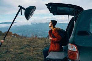 woman tourist with a backpack sits on the trunk of a car in the mountains in autumn in nature photo