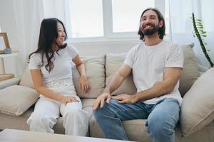 A man and a woman sitting at home on the couch in white stylish t-shirts and chatting merrily smiling and laughing at home. Male and female friendship photo