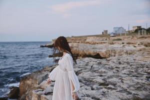 un mujer camina en un blanco vestir en el rocas por el Oceano foto