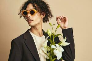 Stylish handsome tanned curly man in trendy sunglasses hold lilies posing isolated on over beige pastel background. Fashion New Collection offer. Retro style concept. Free place for ad photo