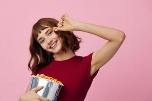 Smiling cheerful pretty cute redhead lady in red shirt with popcorn ready to watch movie posing isolated on over pink studio background. Copy space Banner. Fashion Cinema concept. Entertainment offer photo