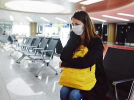 mujer vistiendo un médico máscara a el aeropuerto con un amarillo mochila esperando para un vuelo foto