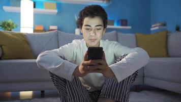 pensativo Adolescente masculino utilizando teléfono, sonriente y mecanografía en teclado. adolescente masculino sentado solo en el piso a hogar mensajes de texto en su teléfono inteligente pensativamente y alegremente video