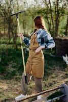 un mujer sonriente hermosamente y mirando a el cámara, un granjero en trabajo ropa y un delantal trabajando al aire libre en naturaleza y participación un rastrillo a reunir césped y forraje para el animales en el jardín foto