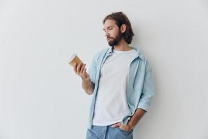 Lanza libre milenario hombre con un barba Bebiendo café desde un reciclado taza en elegante hipster ropa blanco camiseta azul pantalones y camisa en un blanco antecedentes foto