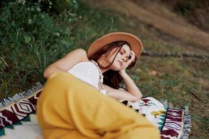 A woman in eco-friendly clothing in a hippie look is lying on a colored plaid smiling and looking at an autumn sunset in nature in the park. Lifestyle on a journey of harmony with the world photo