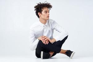 a guy in trousers in sneakers and a shirt sits on the floor and gestures with his hands in full growth photo