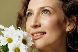 Close up portrait of smiling curly female in linen casual shirt hold chamomiles flowers near face looks up posing isolated on over olive green pastel background. Nature is beauty concept. Copy space photo