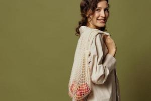 Smiling curly cute female in linen casual shirt hold string bag with fruit behind her back looks at camera posing isolated on olive green background. Eco-friendly Natural products concept. Copy space photo