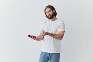 A man with a beard blogger in a white T-shirt with a phone and wireless headphones looks into the phone and spreads his hands to the side on a white background isolated copy space photo