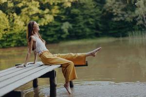 A young happy hippie woman sitting on the lake shore on a bridge in eco clothing made of natural materials in harmony with nature in the fall photo