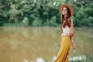 un joven mujer en un hippie Mira viajes en naturaleza por el lago vistiendo un sombrero y amarillo pantalones en el otoño foto