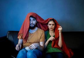 a woman and a man under a red blanket on the couch watching tv in the evening photo