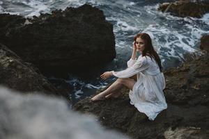 bonito mujer con mojado pelo en un blanco vestir en el costa se sienta en el rocas foto